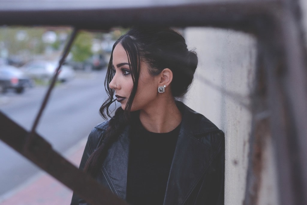 A dark-haired woman wearing an all-black ensemble showcases chunky onyx stud earrings while leaning against a wall near a chain link fence