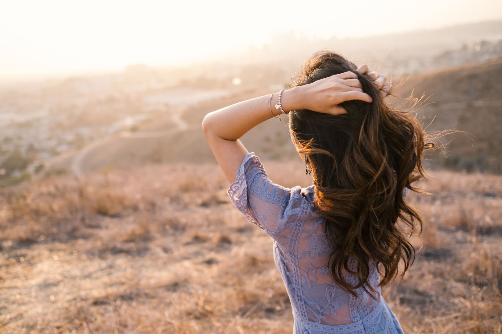 Facing away from the camera, a woman runs her hands through her hair while showing off a heart pendant and gemstone pendant