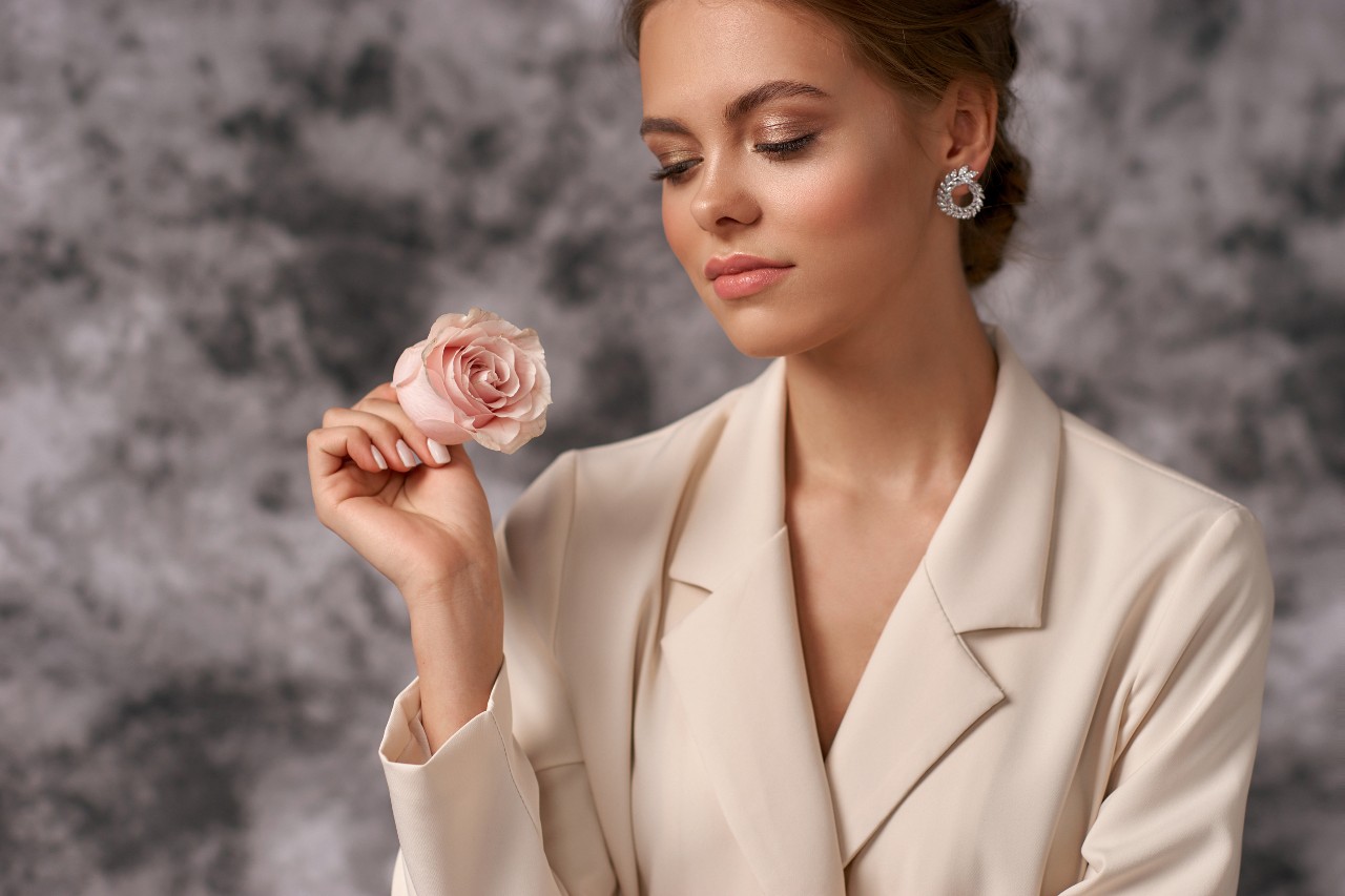 A woman in a tan blazer and diamond earrings holds a pink rose.