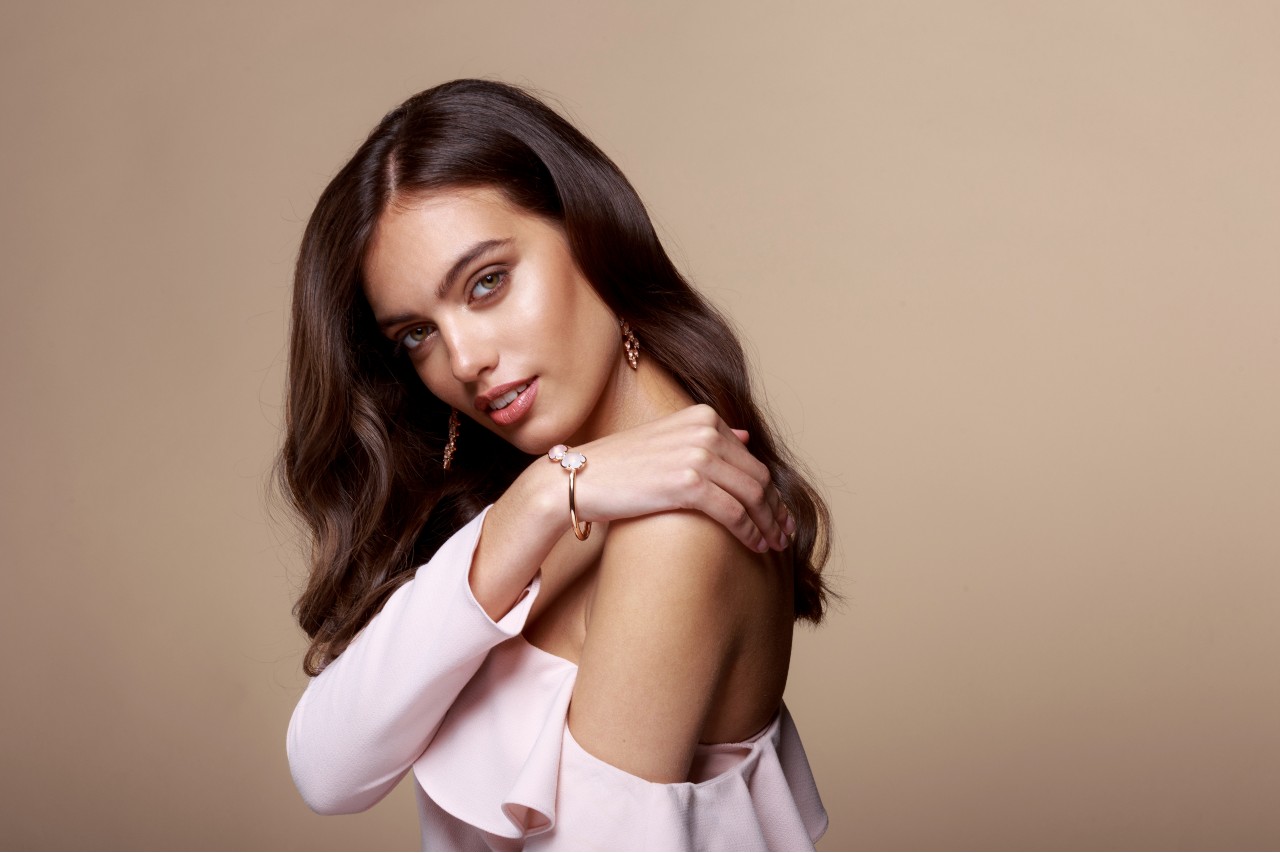 A woman in a pink off-the-shoulder top wears diamond jewelry and poses.