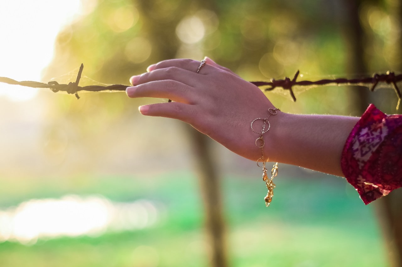 A close-up of a woman’s hand adorned with a gold bracelet, gently touching a barbed wire fence.