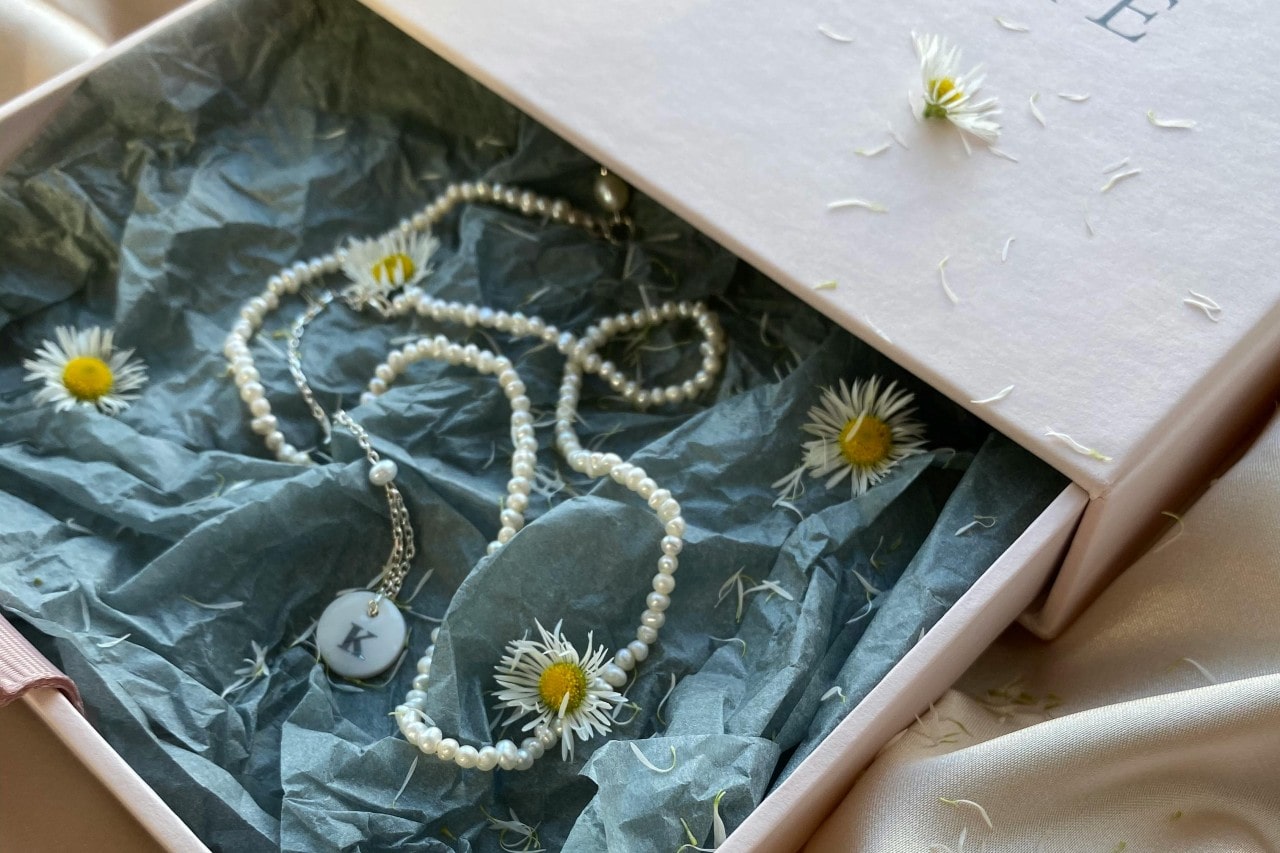 A pearl necklace with a monogram pendant displayed. in a gift box with tissue paper and daisies.