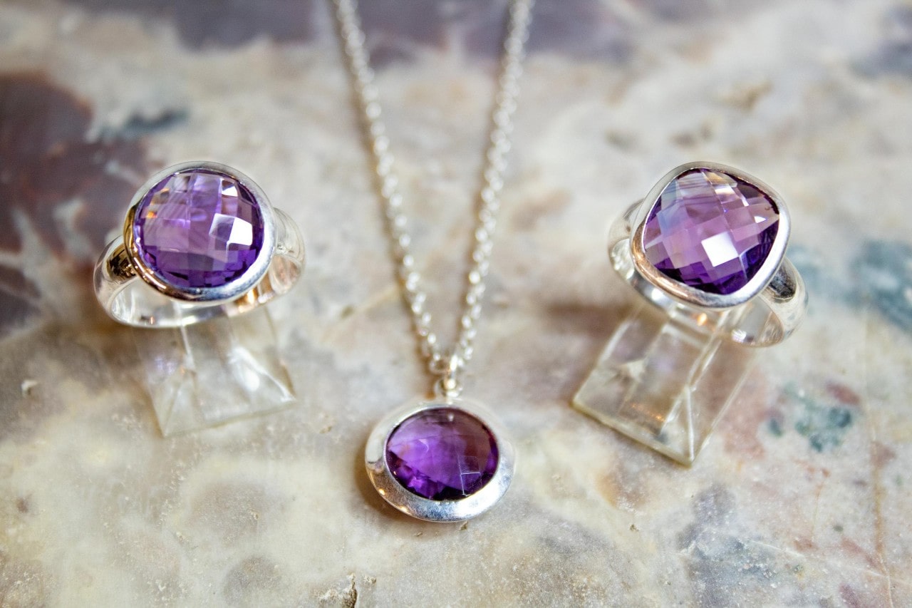 A close-up of a matching set of two amethyst rings and an amethyst necklace on a quartz backdrop.