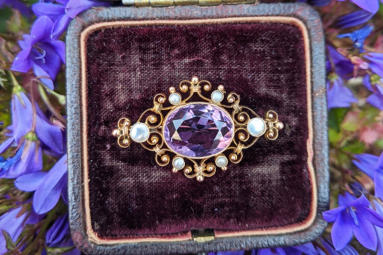 A close-up of an ornate amethyst brooch displayed in a velvet box, surrounded by purple flowers.