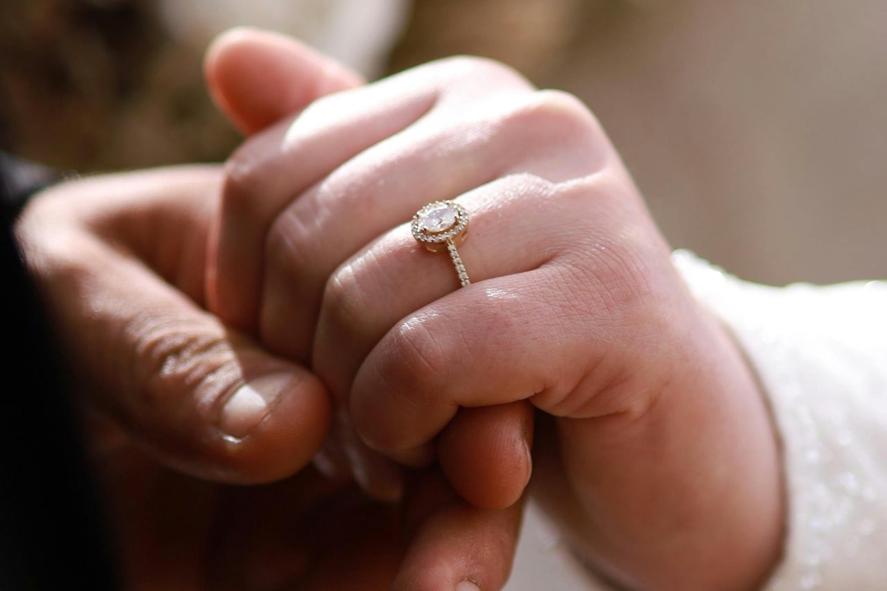 a man’s hand holding a woman’s that is adorned with a yellow gold halo engagement ring