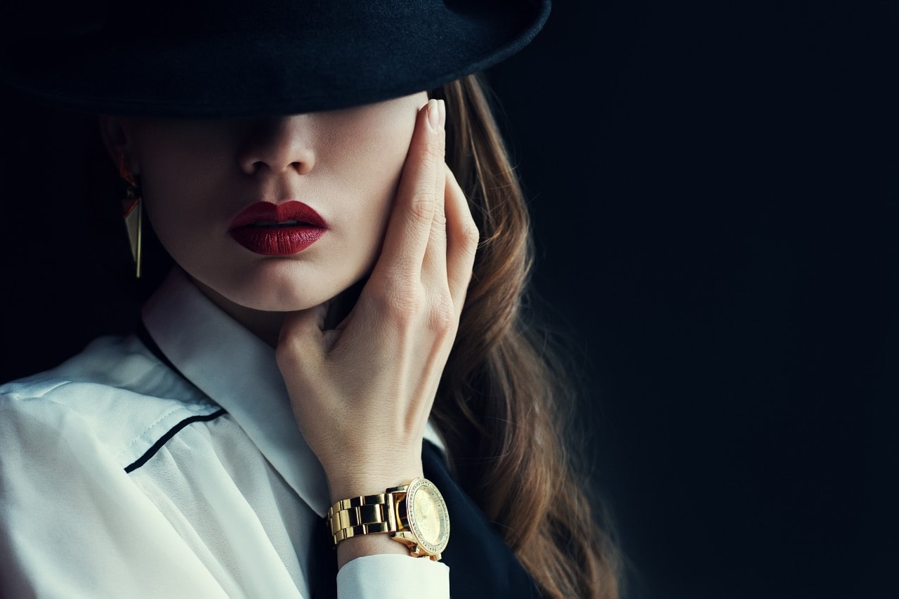 A close-up of a stylish woman whose face is covered by a hat, wearing a luxury watch and distinctive pair of earrings.