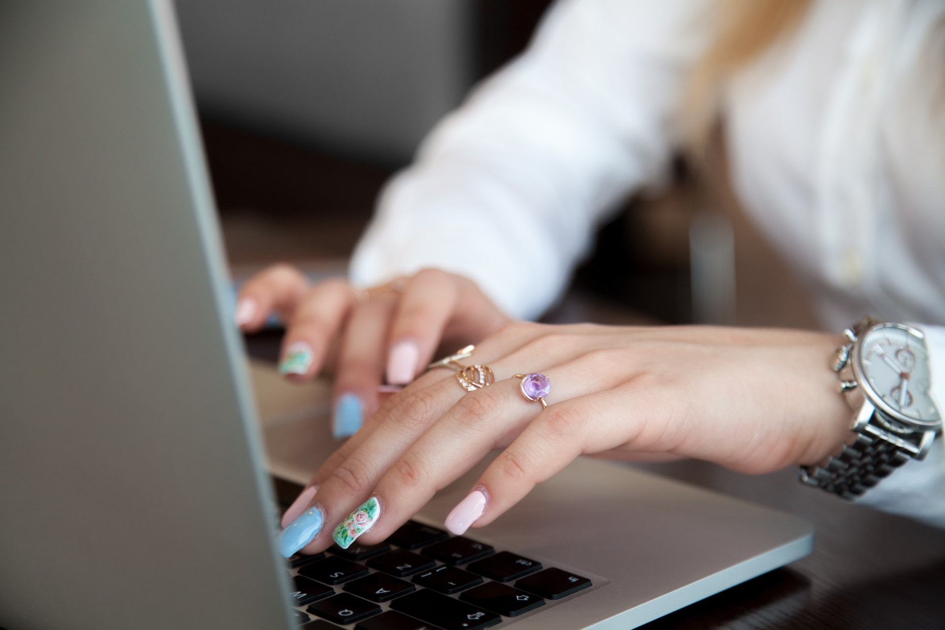 A close-up of a woman’s manicured hands typing on a laptop, her fingers adorned with multiple rings, and a luxury watch on her wrist.