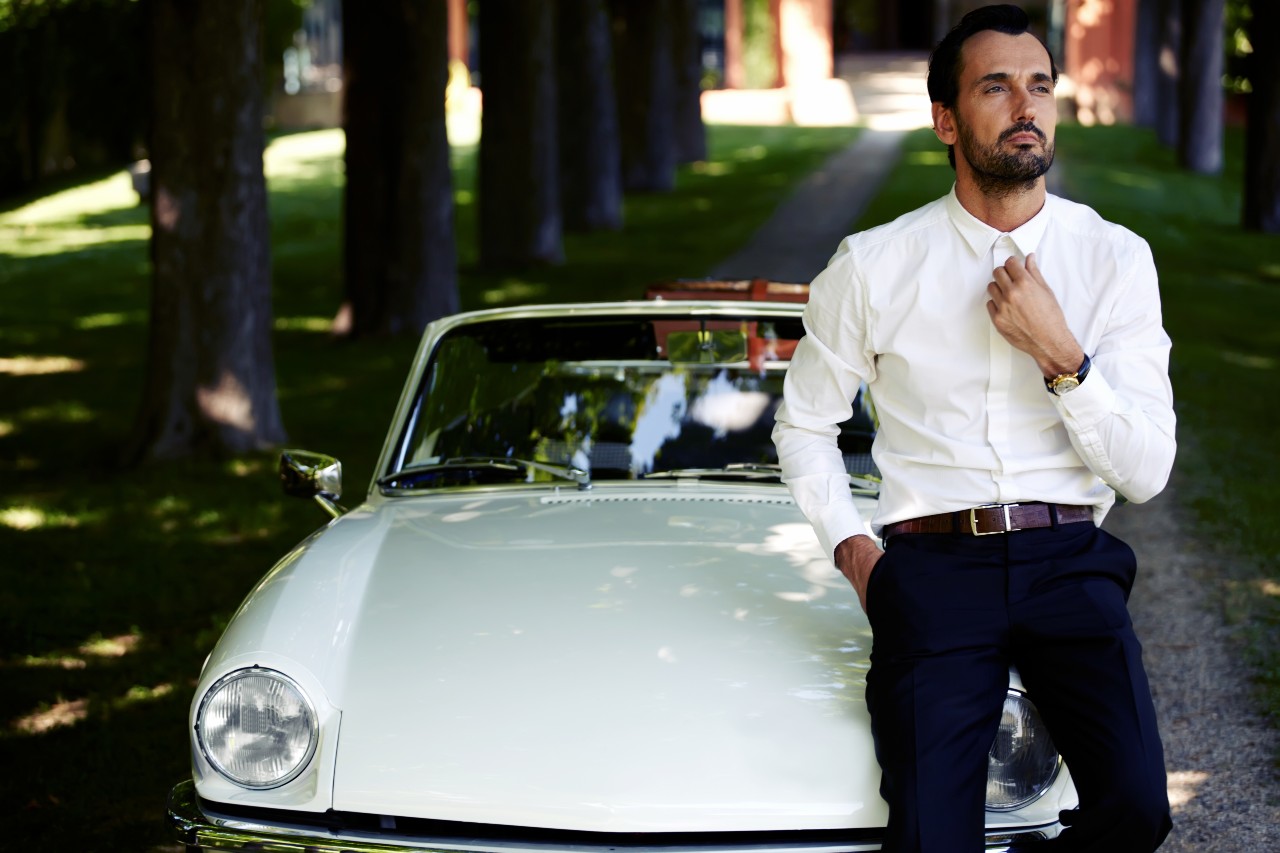 A shot of a well-dressed man in a luxury watch posing before a vintage sports car.