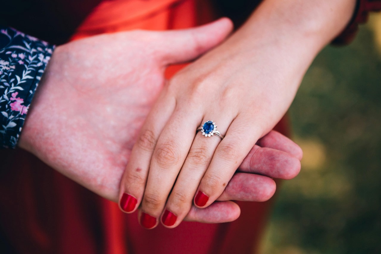 A close-up of a bride-to-be’s hand in her groom-to-be’s, adorned with a sapphire engagement ring.