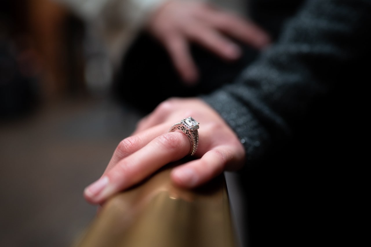 A close-up of a woman’s hand, adorned with an opulent engagement ring, resting on a wooden rail.