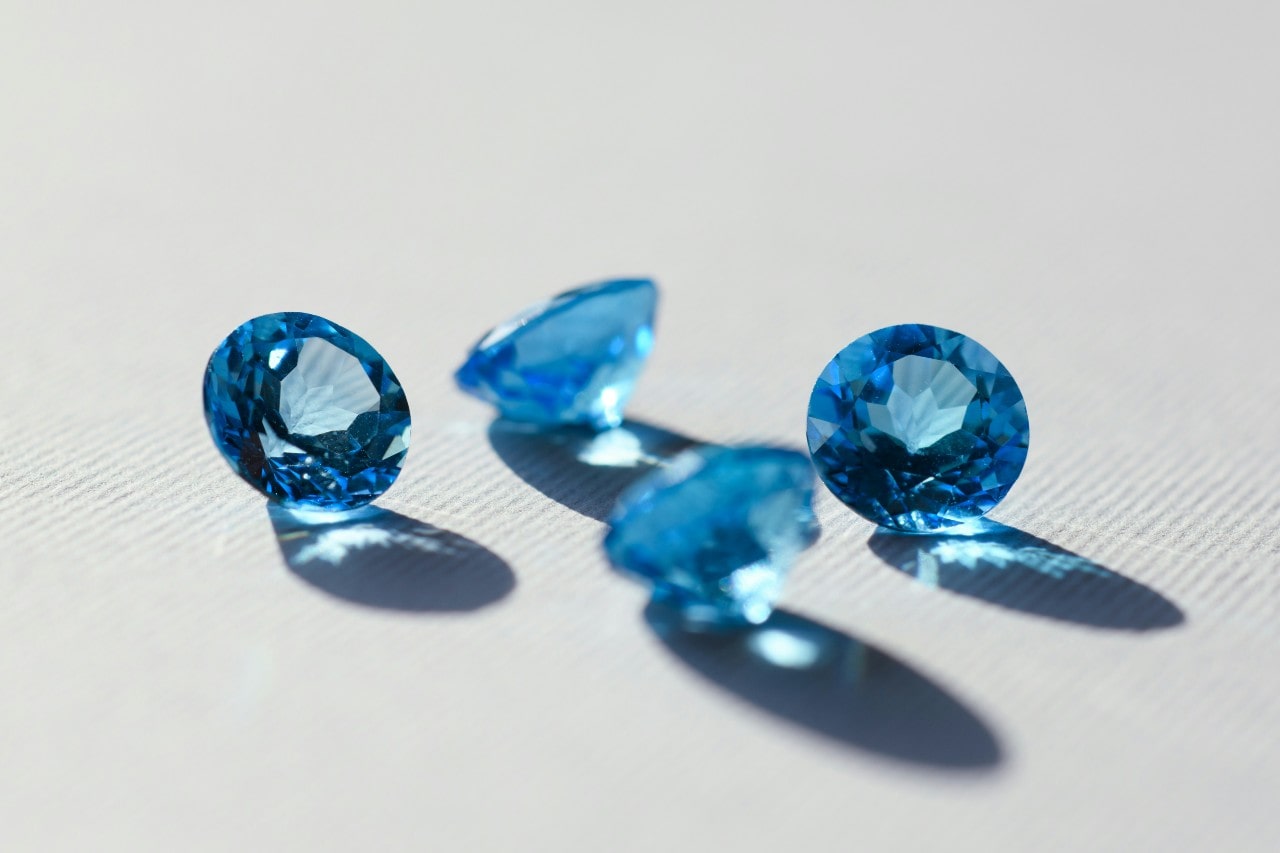 Three vivid blue faceted gemstones rest on a white surface, illuminated by natural light.