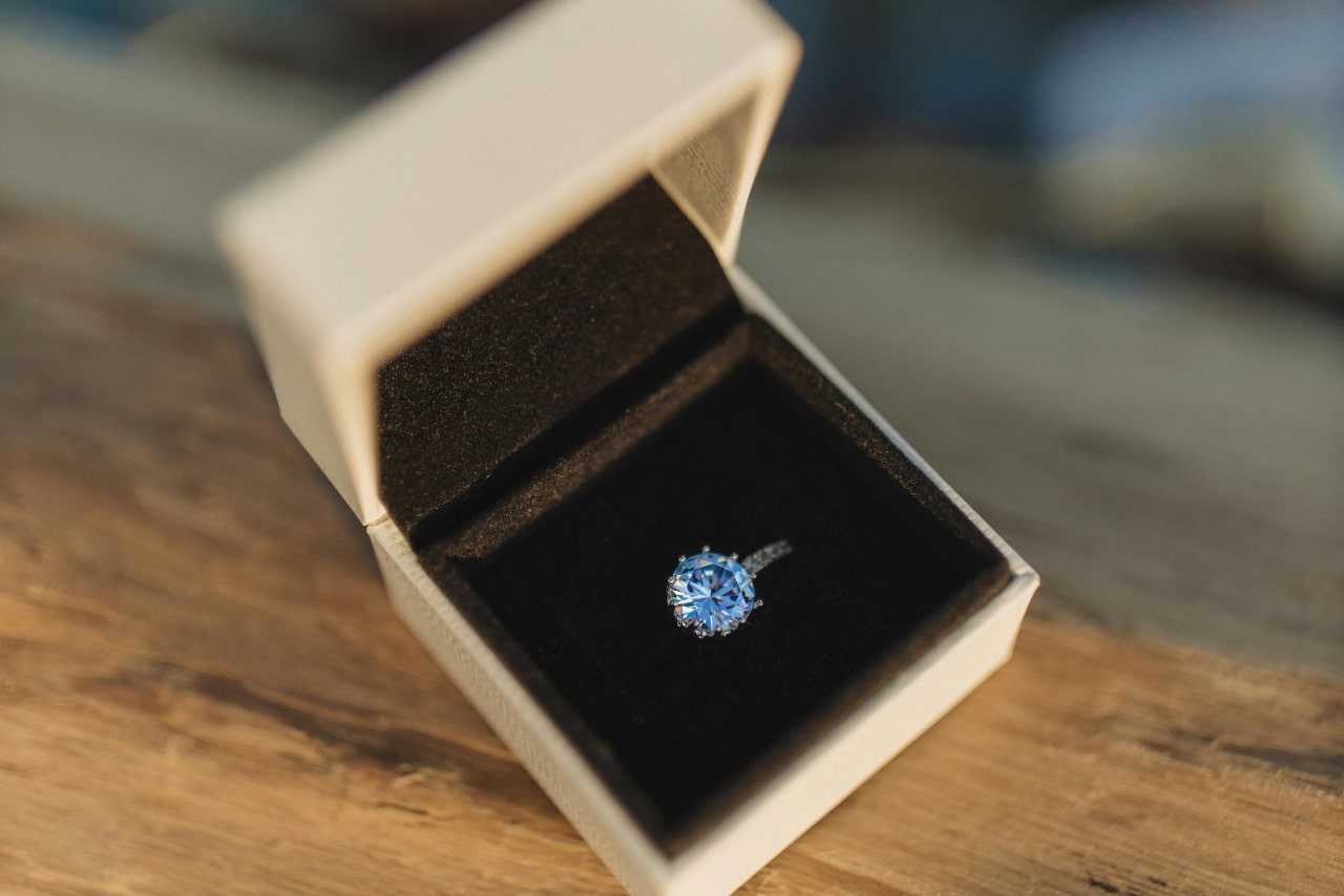 A sparkling round-cut sapphire engagement ring is displayed inside an open white ring box lined with black velvet.