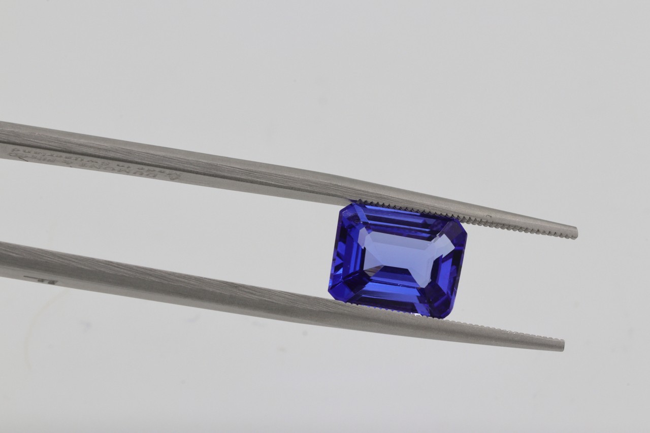 An emerald-cut blue gemstone is securely held in a pair of jeweler’s tweezers against a clean white background.