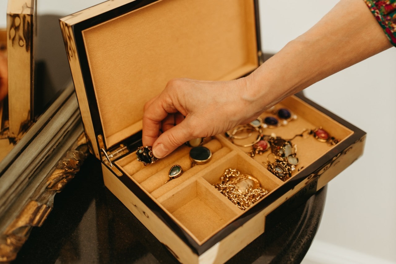 A hand reaches into an open jewelry box filled with rings, necklaces, and gemstone jewelry.